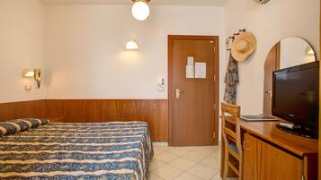 A hotel room with a bed, a desk with a television, a chair, a wooden wardrobe, a door with a calendar, and a ceiling fan.