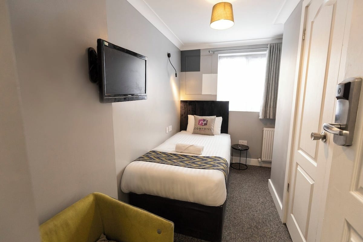 Een hotelkamer met een bed, een aan de muur bevestigde televisie, een nachtkastje, een stoel en een raam met gordijnen.