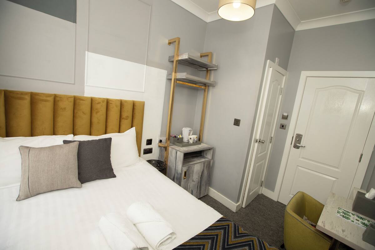 Een hotelkamer met een groot bed, een houten hoofdbord, een klein tafeltje met een waterkoker en een prullenbak.