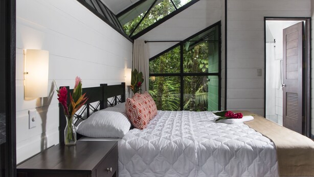 Un dormitorio con cama, mesita de noche, lámpara y ventana con vistas al verdor.