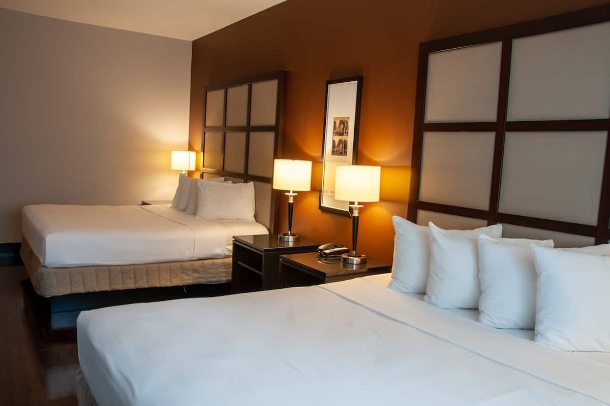 A hotel room with two beds, each with white linens and pillows, a nightstand with a lamp, and a framed picture on the wall.