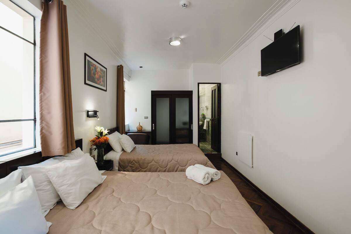 A hotel room with two beds, a TV, a window with curtains, and a door leading to a balcony.