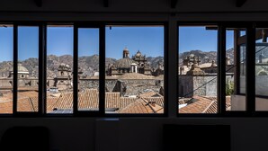 Vue depuis l’intérieur d’un bâtiment, à travers de grandes fenêtres, donnant sur un paysage urbain historique avec des bâtiments à coupoles et des toits en tuiles.