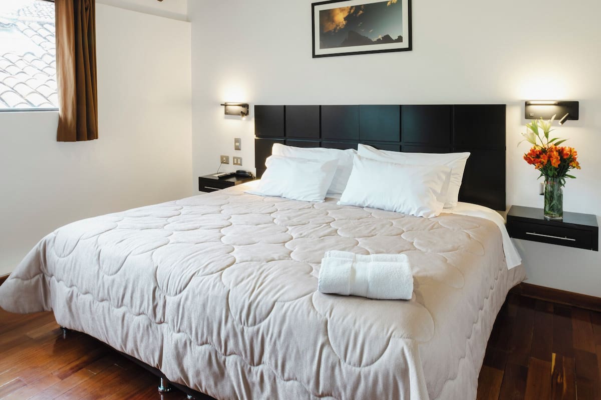 A bedroom with a large bed, wooden beams, a nightstand, a wall-mounted lamp, and a window with curtains.