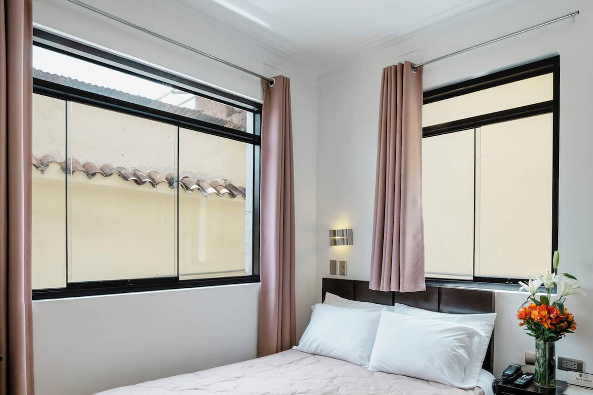 A hotel room with a bed, a nightstand, a vase with flowers, and a window with curtains.