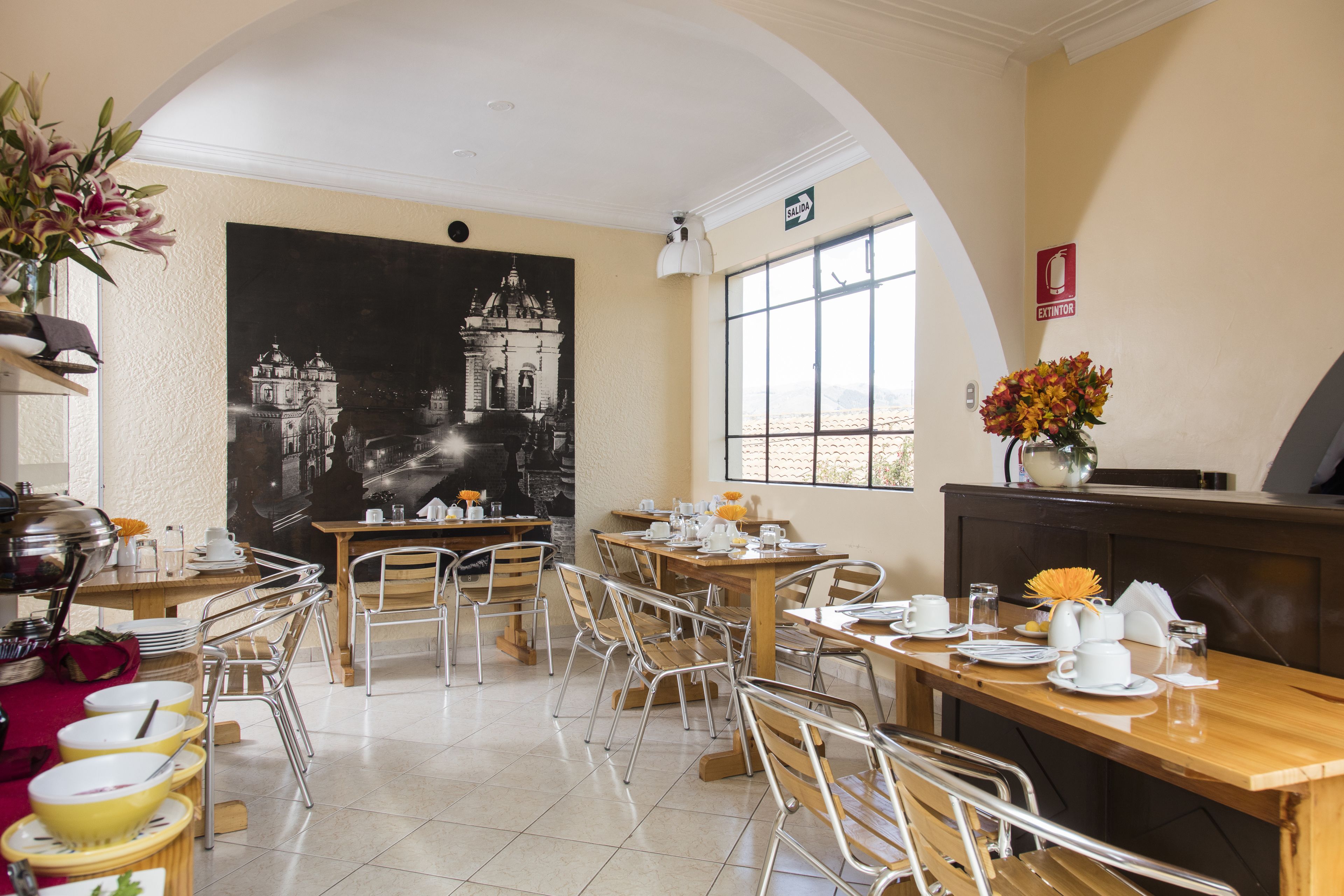 Un comedor con varias mesas y sillas, un gran mural en blanco y negro en la pared, y una ventana con luz natural.