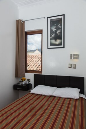 Ein Hotelzimmer mit Bett, einem Fenster mit Vorhängen, einem gerahmten Bild und einem Nachttisch mit Blumen.