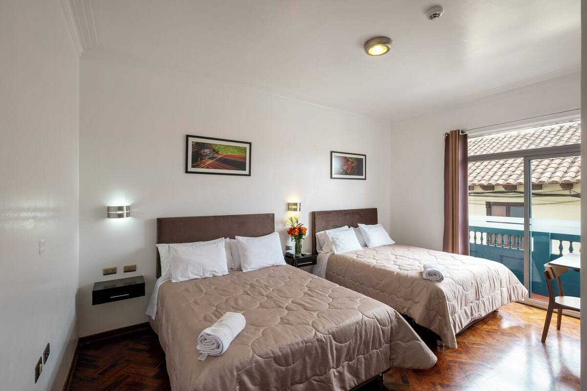 A hotel room with two beds, a nightstand, a small table, and a balcony with a view.