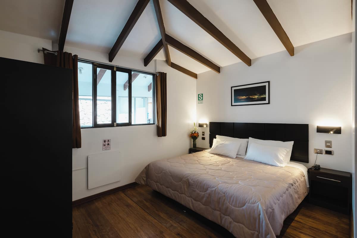 A bedroom with a bed, wooden floor, a window with curtains, a radiator, a nightstand, and a wall-mounted picture.