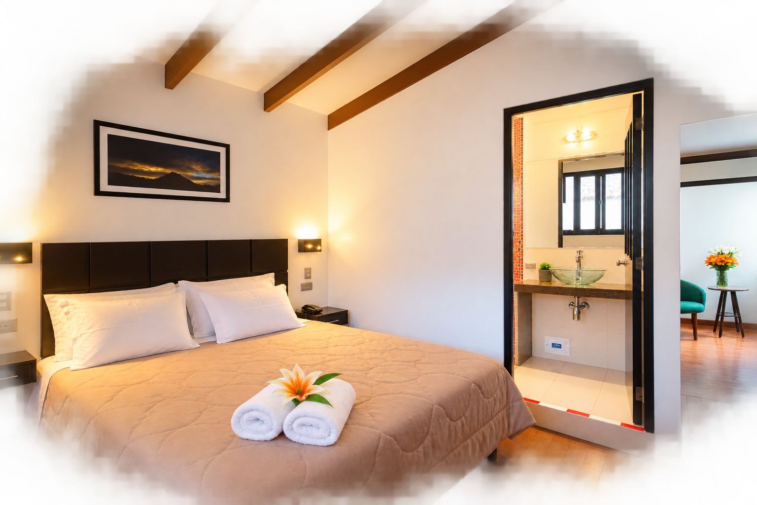 Habitación de hotel con cama, mesita de noche, lámpara de pared, un cuadro enmarcado y baño a la vista a través de una puerta abierta.
