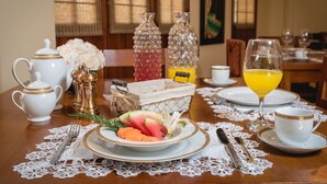 Una mesa de comedor dispuesta con teteras blancas, una tetera con detalles dorados, un juego de salero y pimentero, un frutero con sandía y melón, un vaso de jugo de naranja y una taza blanca con detalles dorados.
