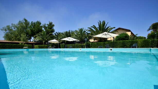 A clear blue swimming pool with lounge chairs and umbrellas, surrounded by greenery and a building in the background. - Tenuta San Francesco (Trinitapoli)