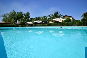 A clear blue swimming pool with lounge chairs and umbrellas, surrounded by greenery and a building in the background.