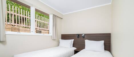Ein Hotelzimmer mit zwei Einzelbetten, einem Holz-Kopfstück, einem Fenster mit Blick auf Grünflächen und einem Nachttisch mit Lampe.