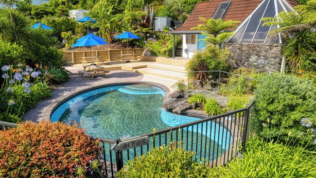 A swimming pool surrounded by lush greenery, with a wooden deck and picnic tables. - The Captains Motel (Russell)