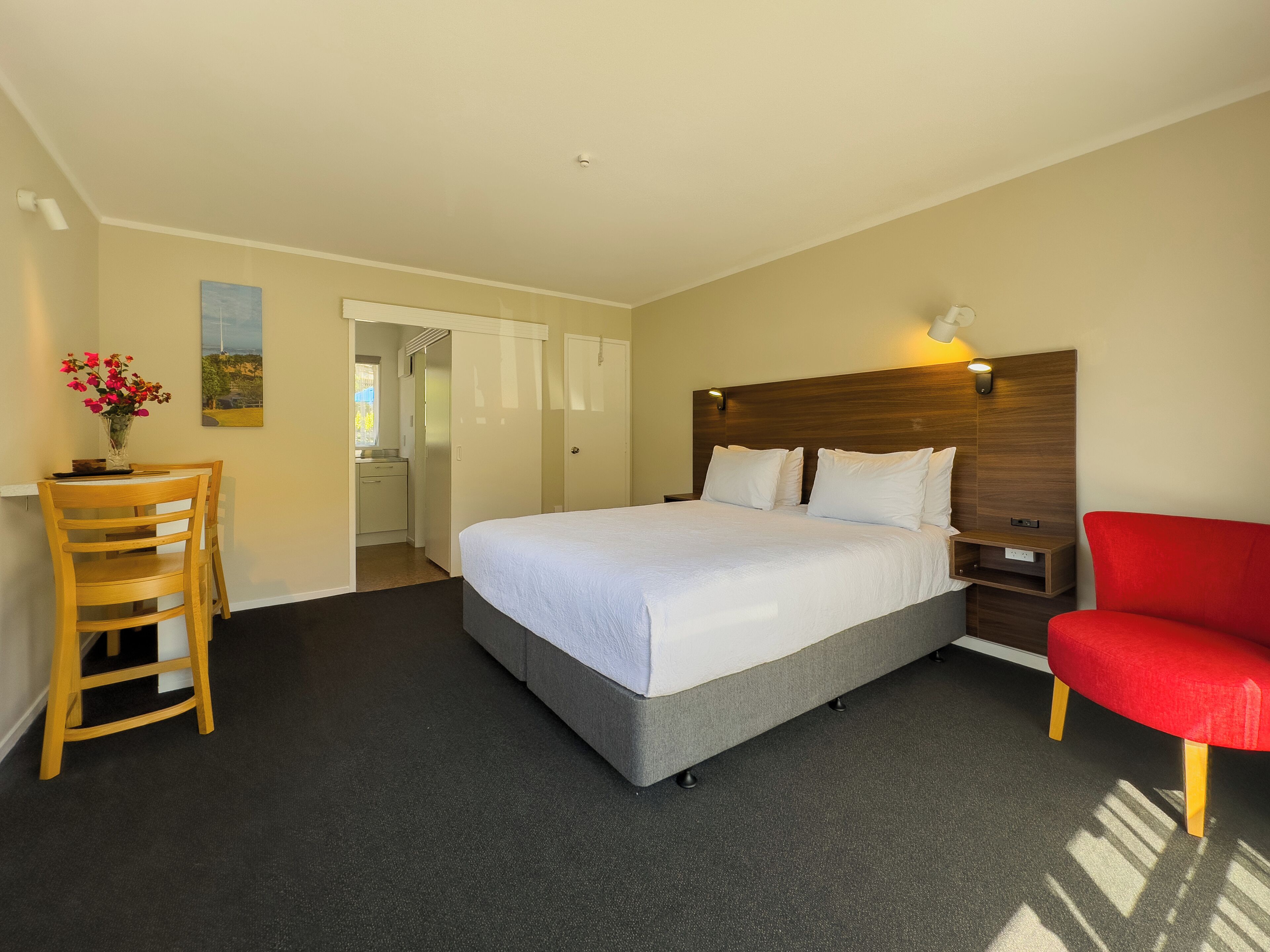 A hotel room with a bed, a desk with a chair, a red chair, a nightstand, and a wall-mounted lamp.