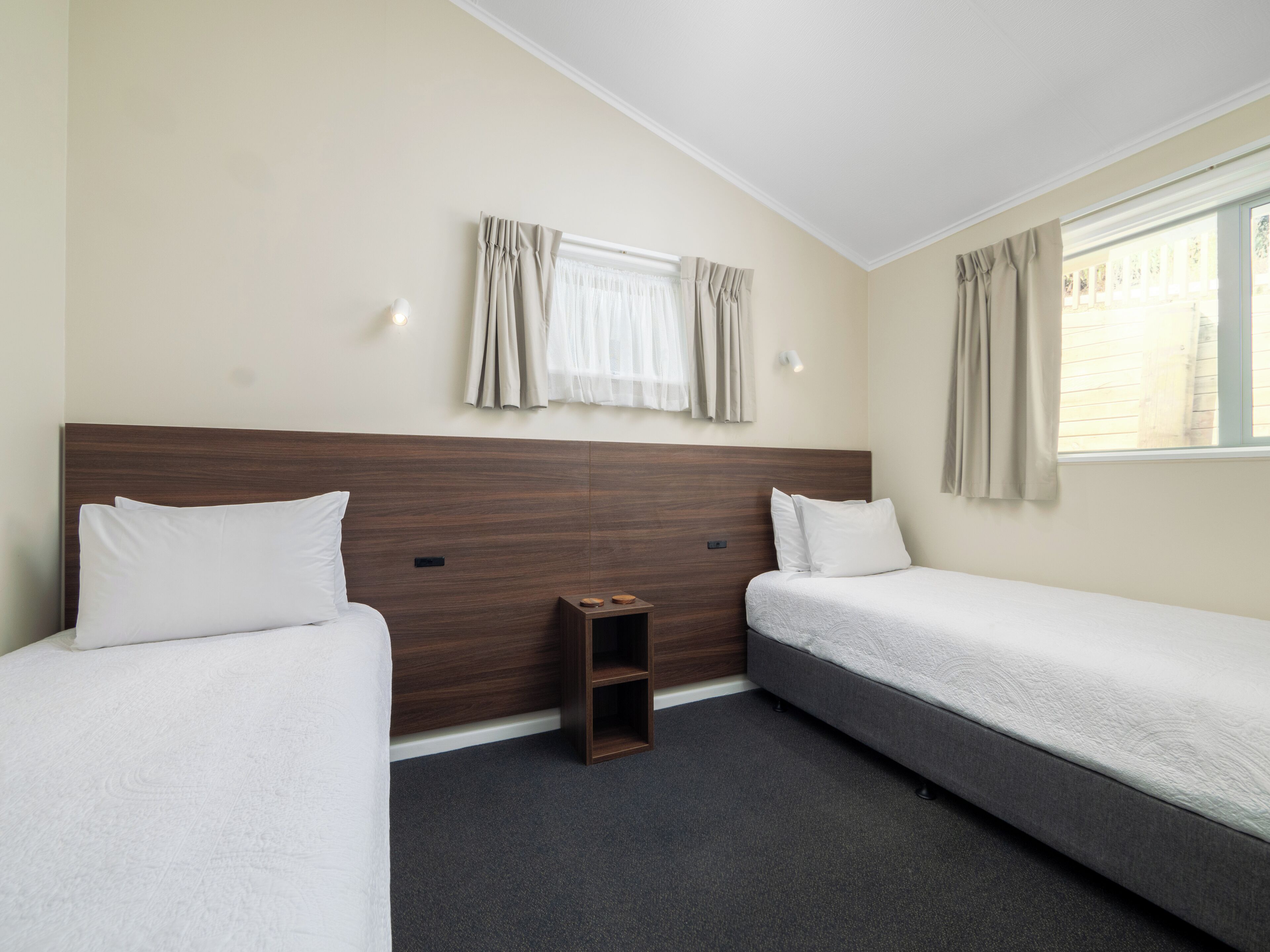 Hotellihuone, jossa on kaksi erillistä sänkyä, pieni puinen yöpöytä, ikkuna, jossa on verhot, ja seinässä oleva valaisin.