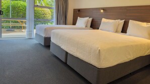 A hotel room with two beds, a large window with curtains, and a view of greenery outside. - The Captains Motel (Russell)