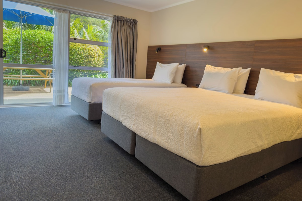 Ett hotellrum med två sängar, ett stort fönster med gardiner och utsikt över grönområden utanför.