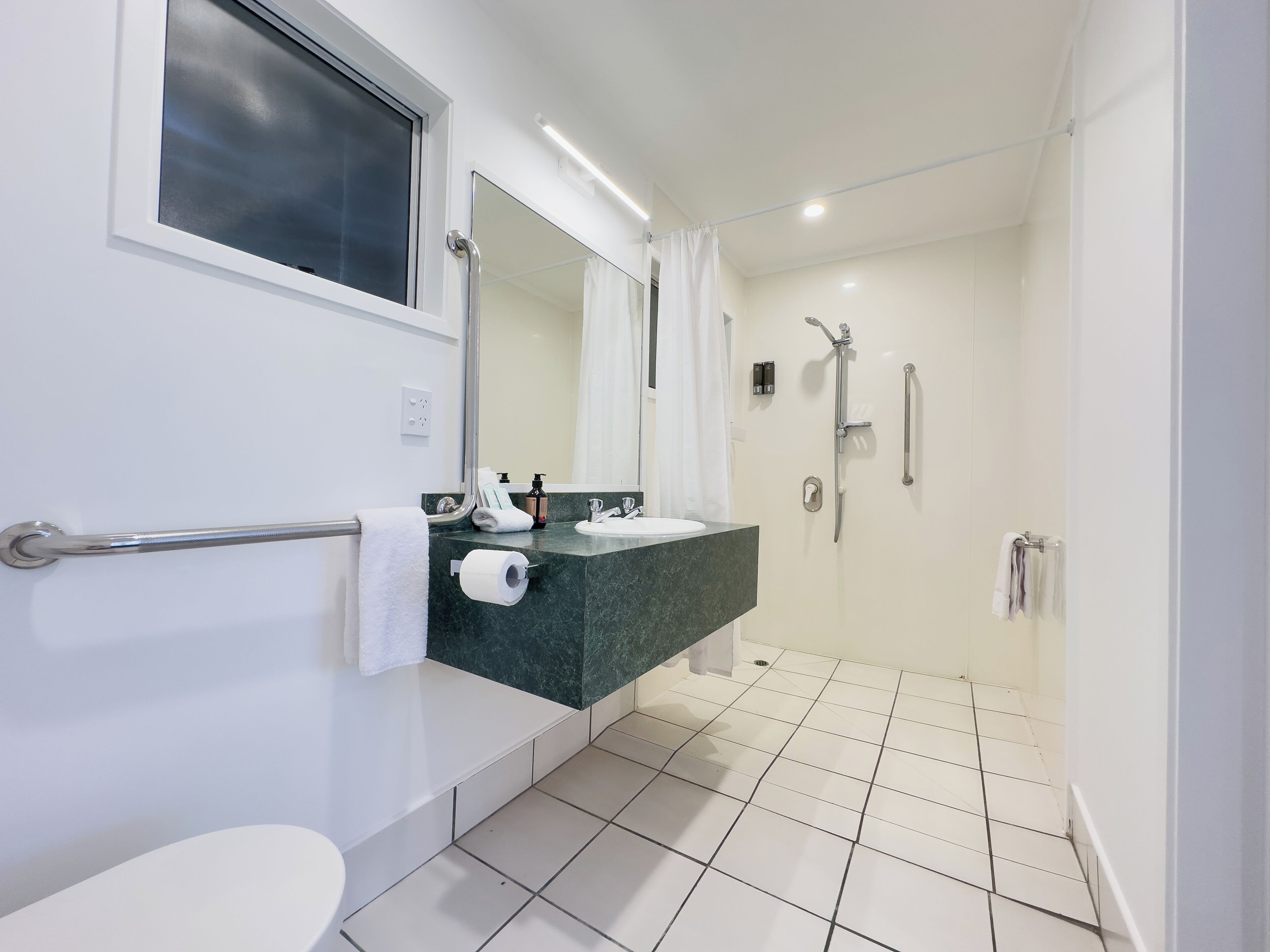 A modern bathroom with a toilet, sink, mirror, shower, and a handrail.