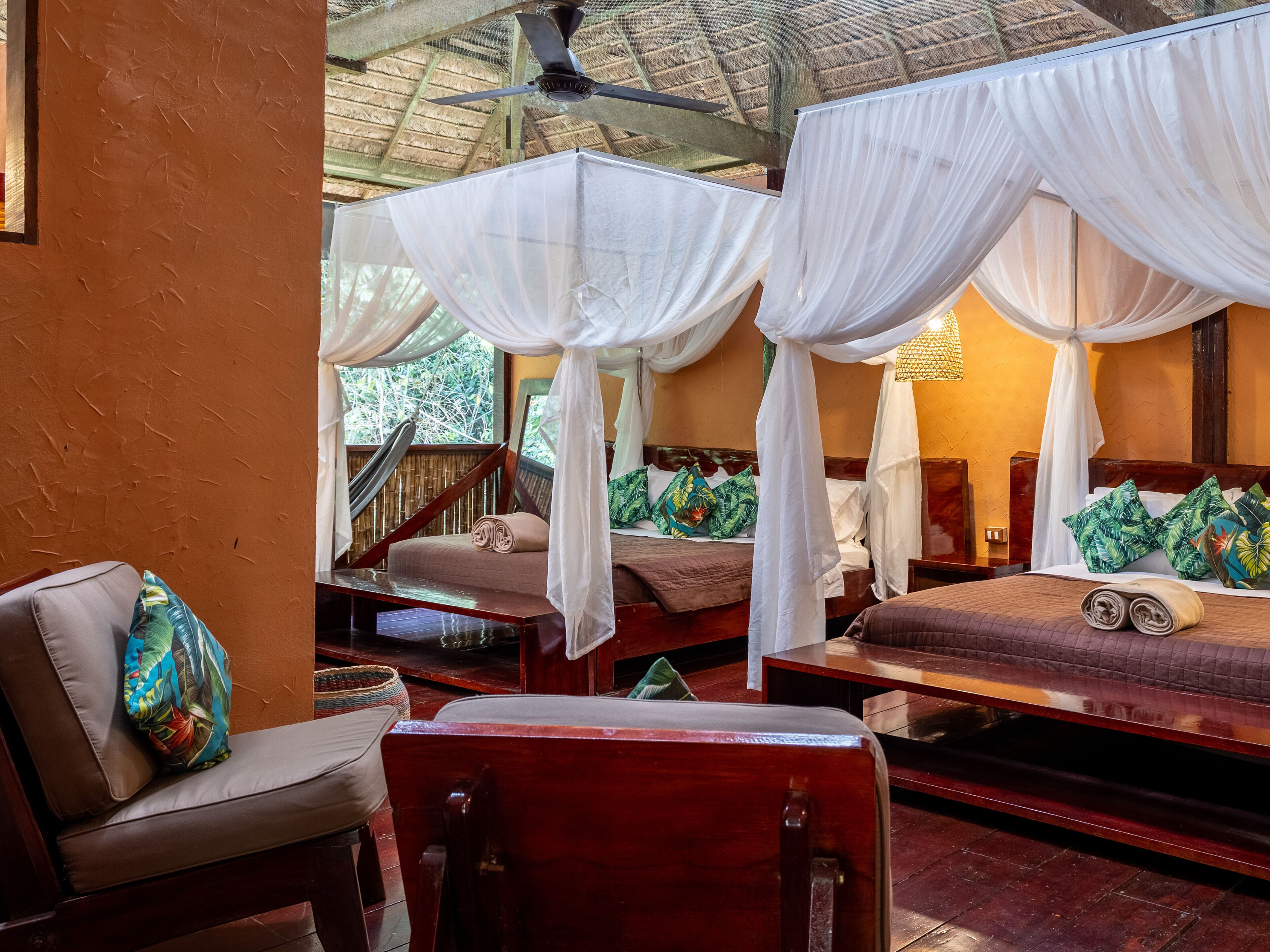 A room with two beds, each with a canopy, a wooden chair, a small table, and a view of the outdoors.