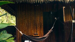 Una hamaca colgando entre dos postes en una habitación con pared de bambú, una mesita con botellas y suelo de madera.