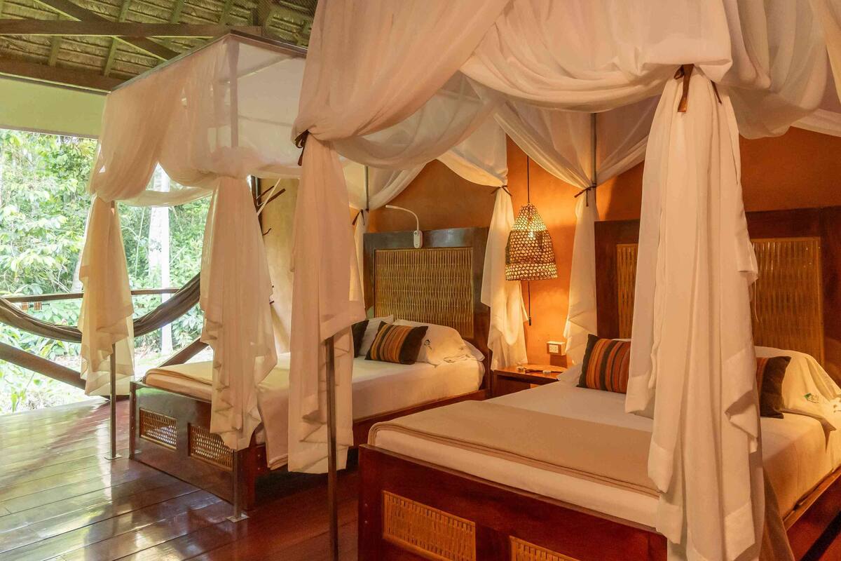 A room with a bed under a canopy, another bed with a headboard, a hanging chair, and a window with a view of greenery.