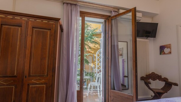 A bedroom with a wooden wardrobe, a bed with a white cover, a television mounted on the wall, and a view of an outdoor patio with a chair and a table. - Giulietta e Romeo (Verona)