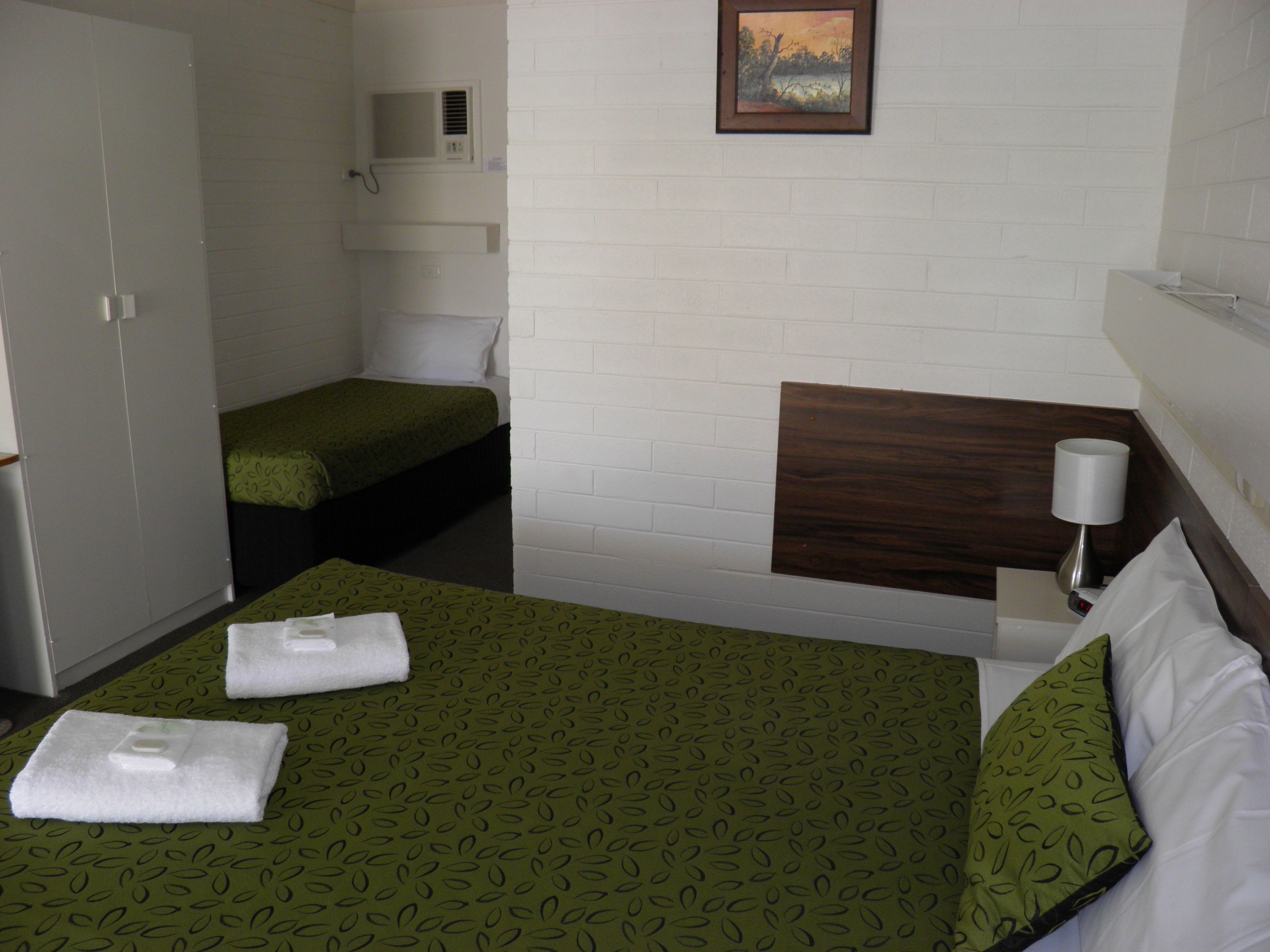 A hotel room with a bed, a bedside table with a lamp, a small bedside table with a phone, a wall-mounted air conditioning unit, and a painting on the wall.