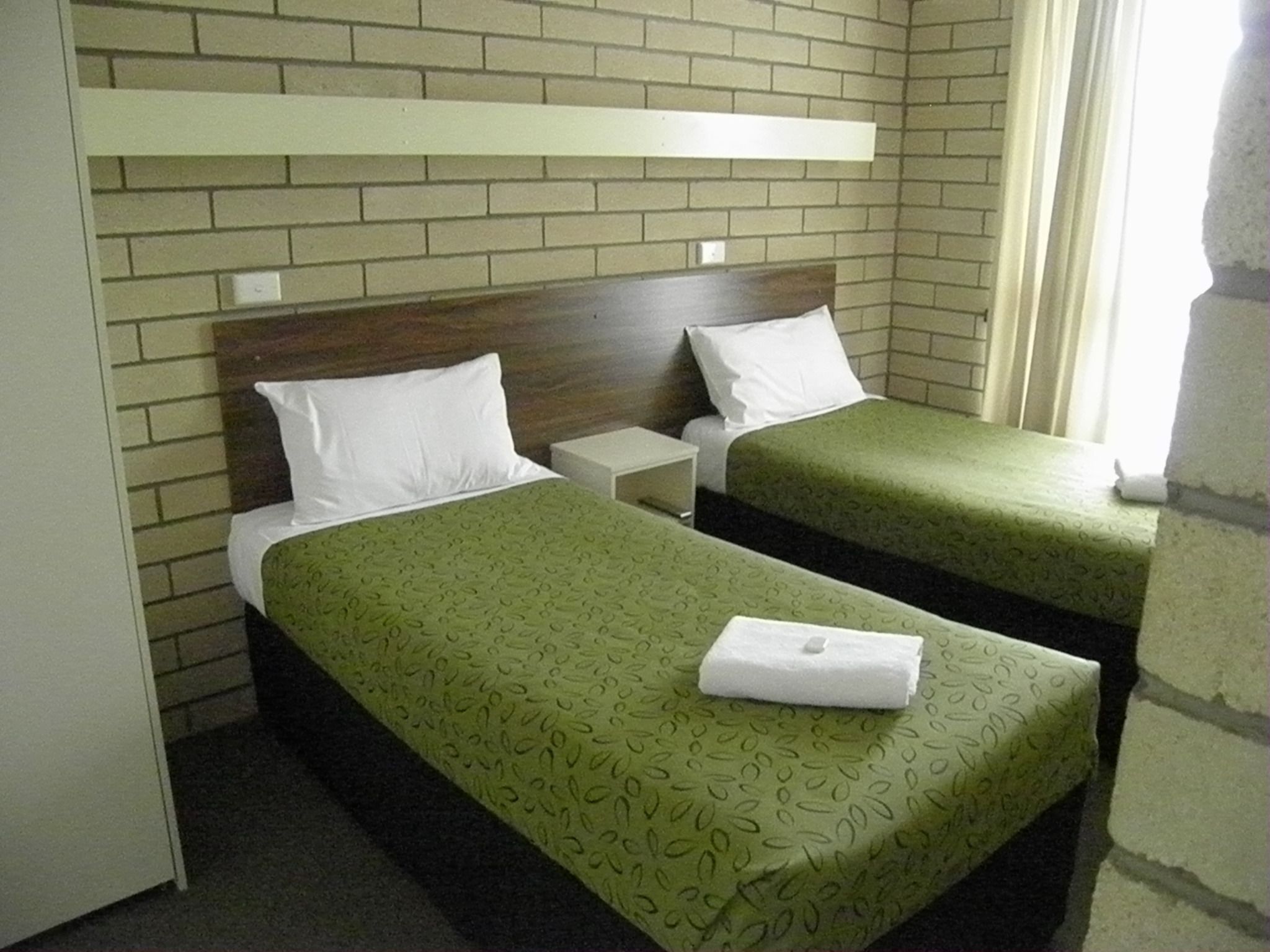 A room with two single beds, each with a green patterned bedspread and white pillows. There is a white nightstand between the beds, and a white shelf above the beds. The room has a brick wall and a window with curtains.