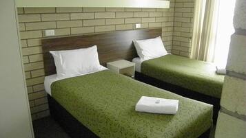 A room with two single beds, each with a green patterned bedspread and white pillows. There is a white nightstand between the beds, and a white shelf above the beds. The room has a brick wall and a window with curtains.