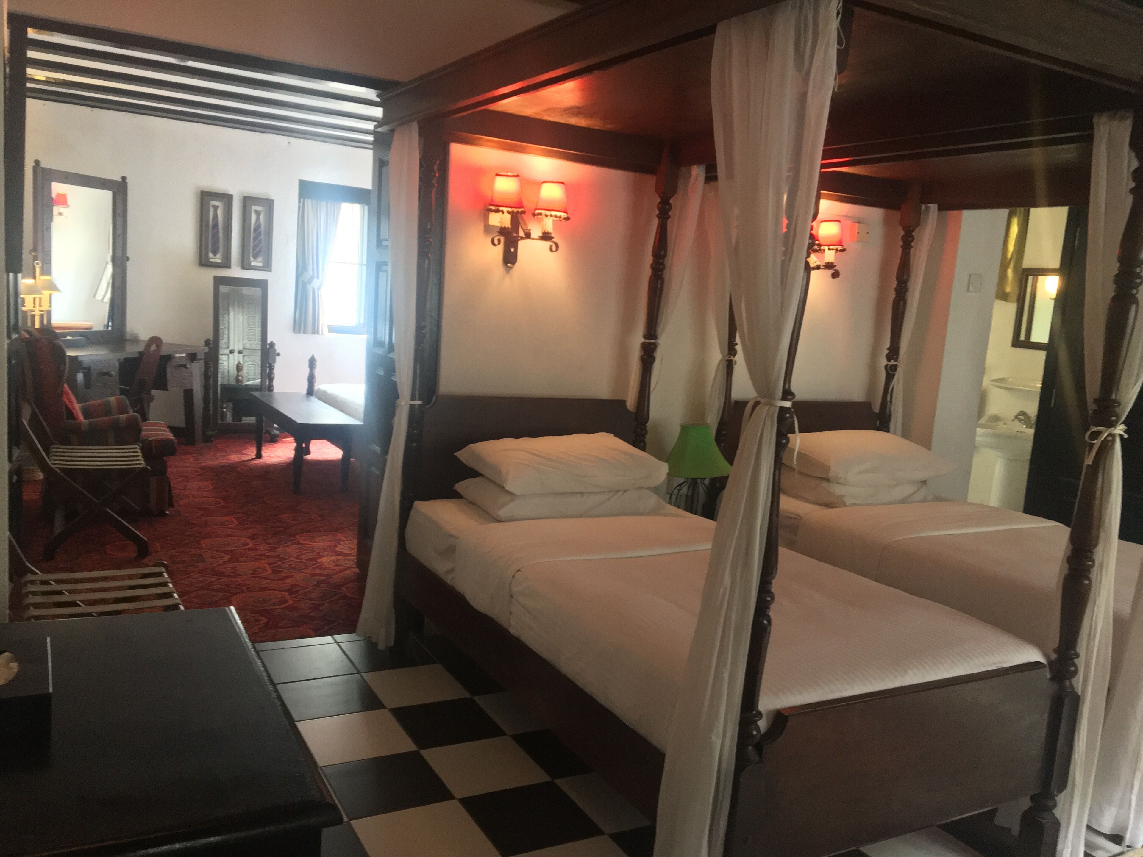 A room with a checkered floor, a four-poster bed with white linens, a desk with a chair, and wall-mounted lamps.