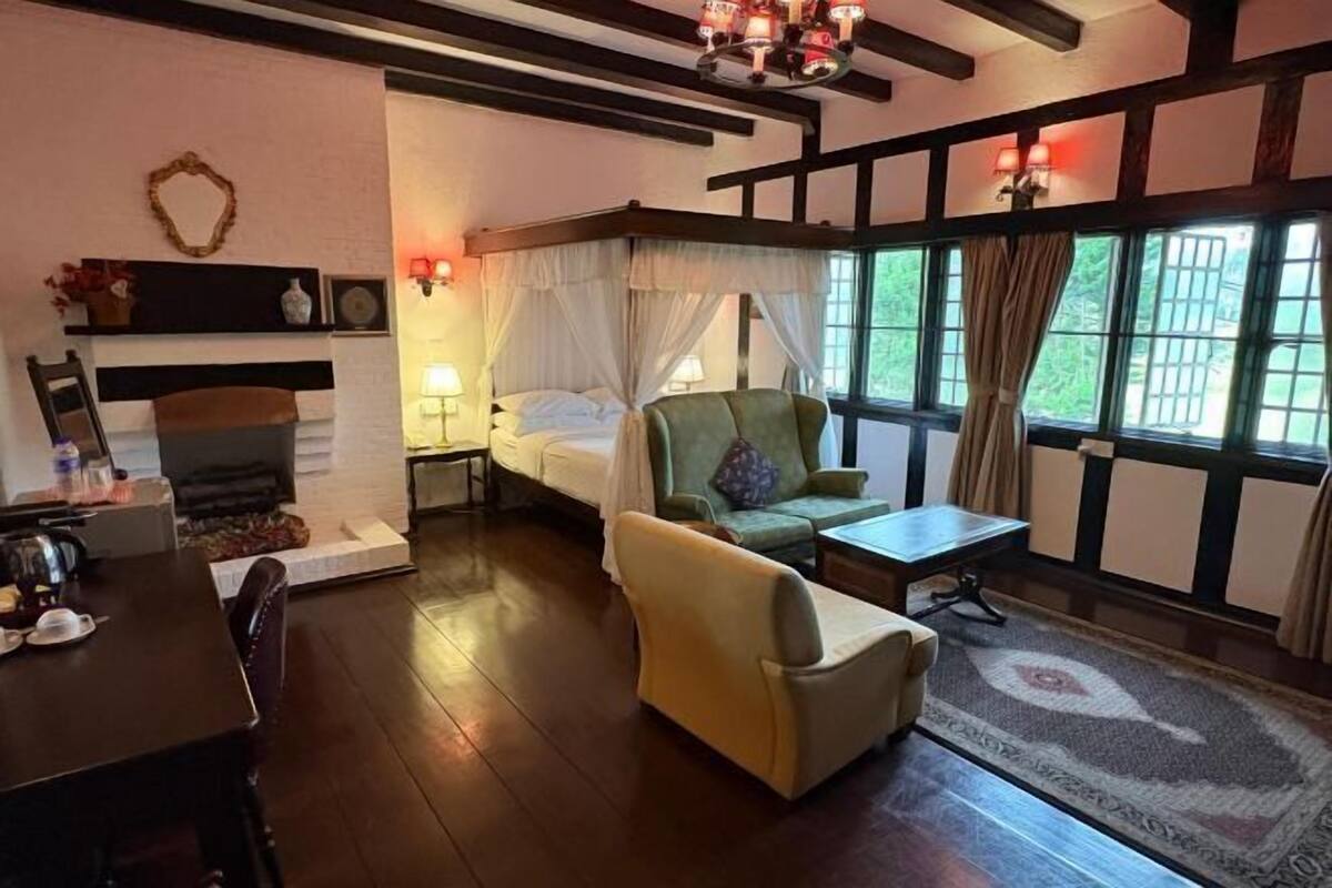Ein Zimmer mit Bett, Sessel, Tisch, Kamin und großen Fenstern.