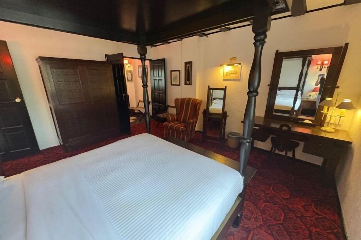 Un lit à baldaquin, un bureau avec une chaise, un miroir et une moquette à motifs rouges dans une chambre d’hôtel.