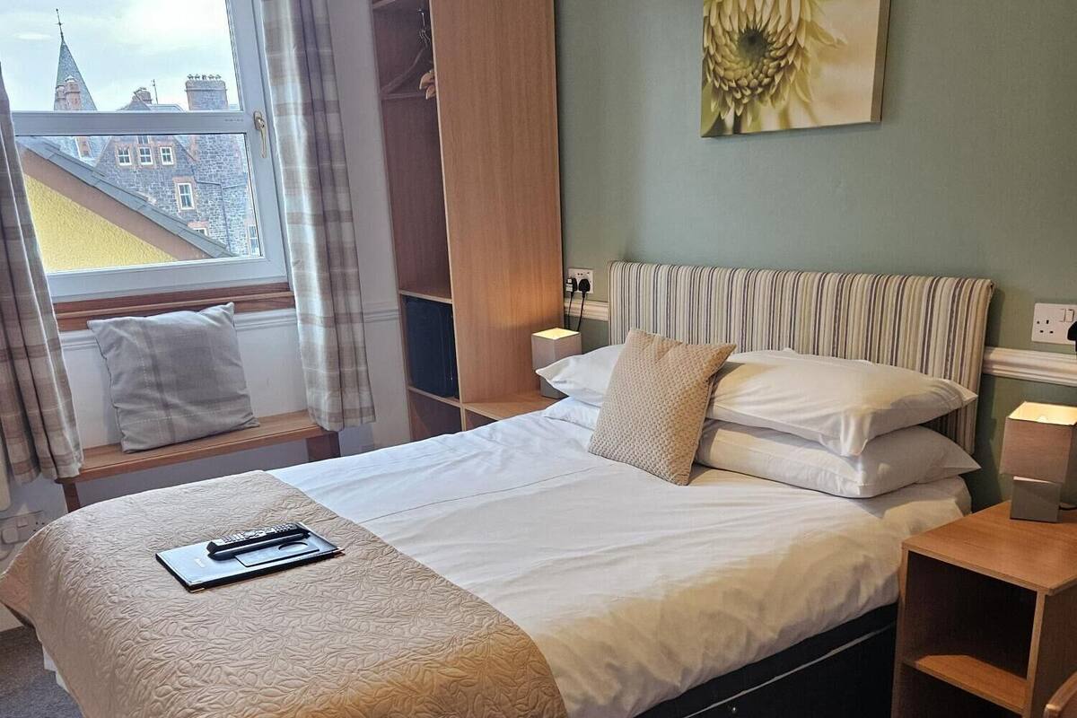 Ein ordentlich eingerichtetes Hotelzimmer mit Bett, Nachttischen, einem Fenster mit Vorhängen, Blick auf Gebäude und einem Bild an der Wand.