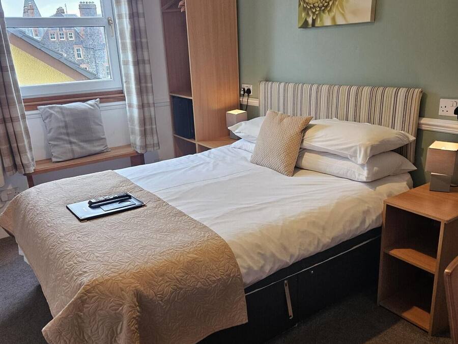 Ein ordentlich eingerichtetes Hotelzimmer mit Bett, Nachttischen, einem Fenster mit Vorhängen, Blick auf Gebäude und einem Bild an der Wand.