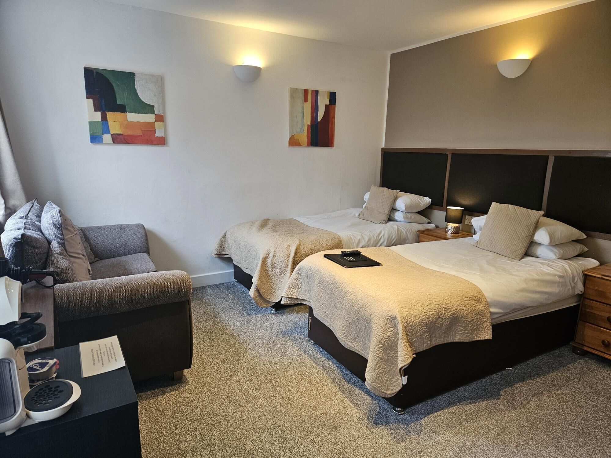A hotel room with two beds, a sofa, a small table with a lamp, and abstract wall art.