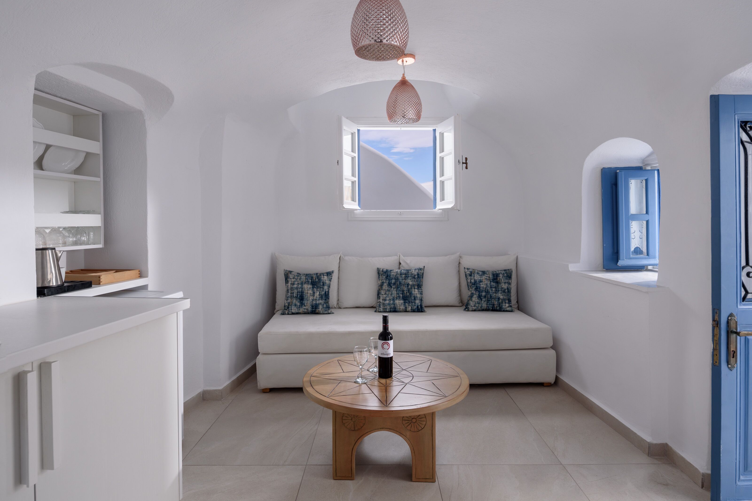Una sala de estar moderna con una mesa de centro redonda de madera, un sofá blanco con cojines estampados, una ventana con vistas y una puerta azul.