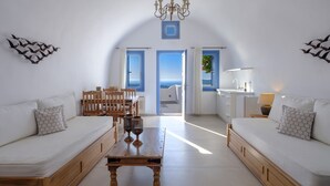 A modern, minimalist room with a wooden dining table, two white sofas, a chandelier, and a view of the sea through the open doors. - Kamares Apartments (Santorini)