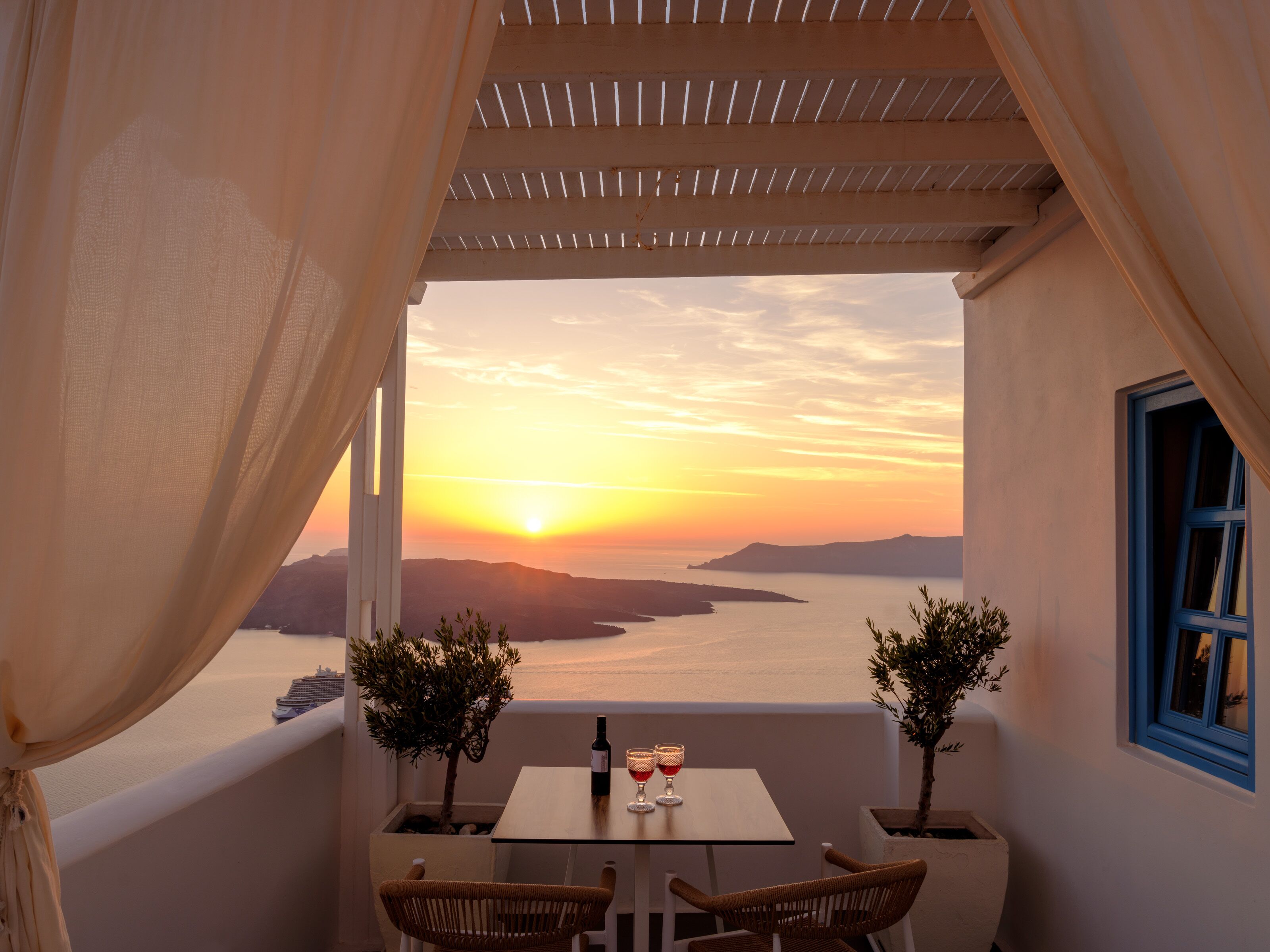 A balcony with a table and chairs, overlooking a sunset over water and hills.