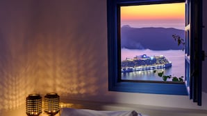 A bedroom with a bed, a nightstand with two decorative lamps, a window with a view of a cityscape at sunset, and a striped curtain. - Kamares Apartments (Santorini)