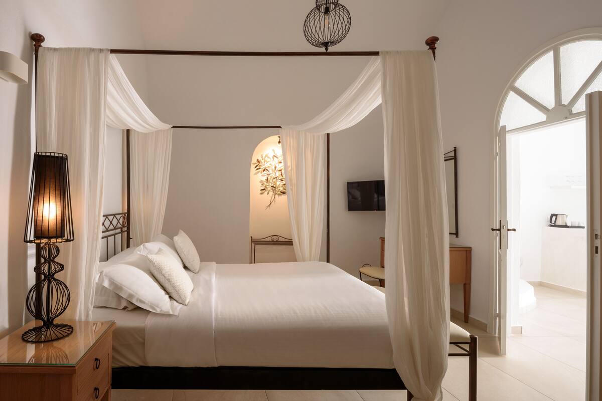 A bedroom with a four-poster bed, a bedside lamp, and a view of the adjacent room.