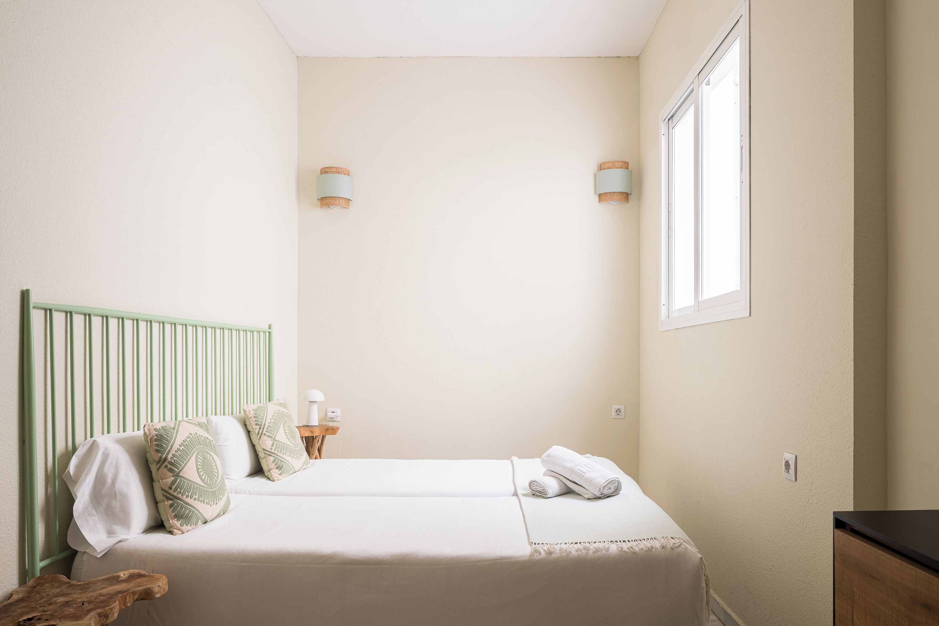 Ein ordentlich bezogenes Bett mit grünem Kopfteil, ein Nachttisch mit Lampe und ein Fenster, das für natürliches Licht sorgt.