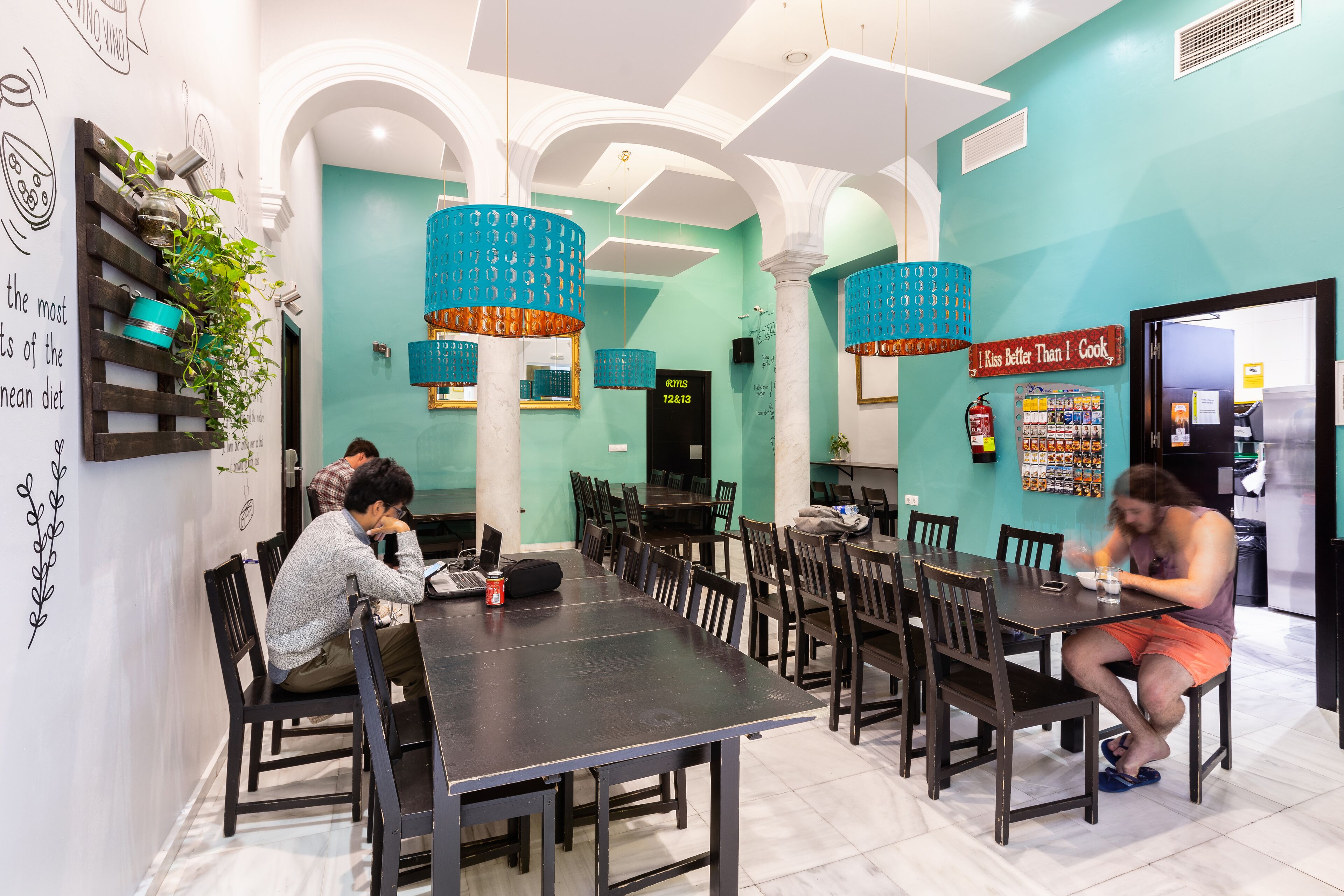 Un comedor con mesas y sillas, una pared con una planta y un letrero, y una pared de color verde azulado.