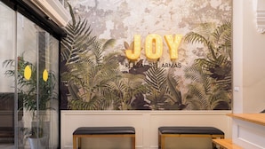 Una zona de recepción con piso a cuadros, dos bancos de madera y un mural con la palabra “JOY” y “PLAZA DE ARMAS”.