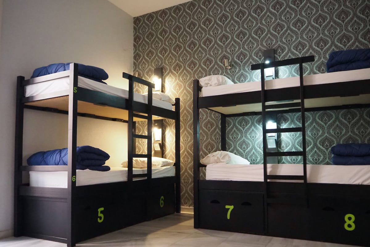 Dos literas con los números 5, 6, 7 y 8, en una habitación con papel tapiz estampado y una mesita de noche con una lámpara.