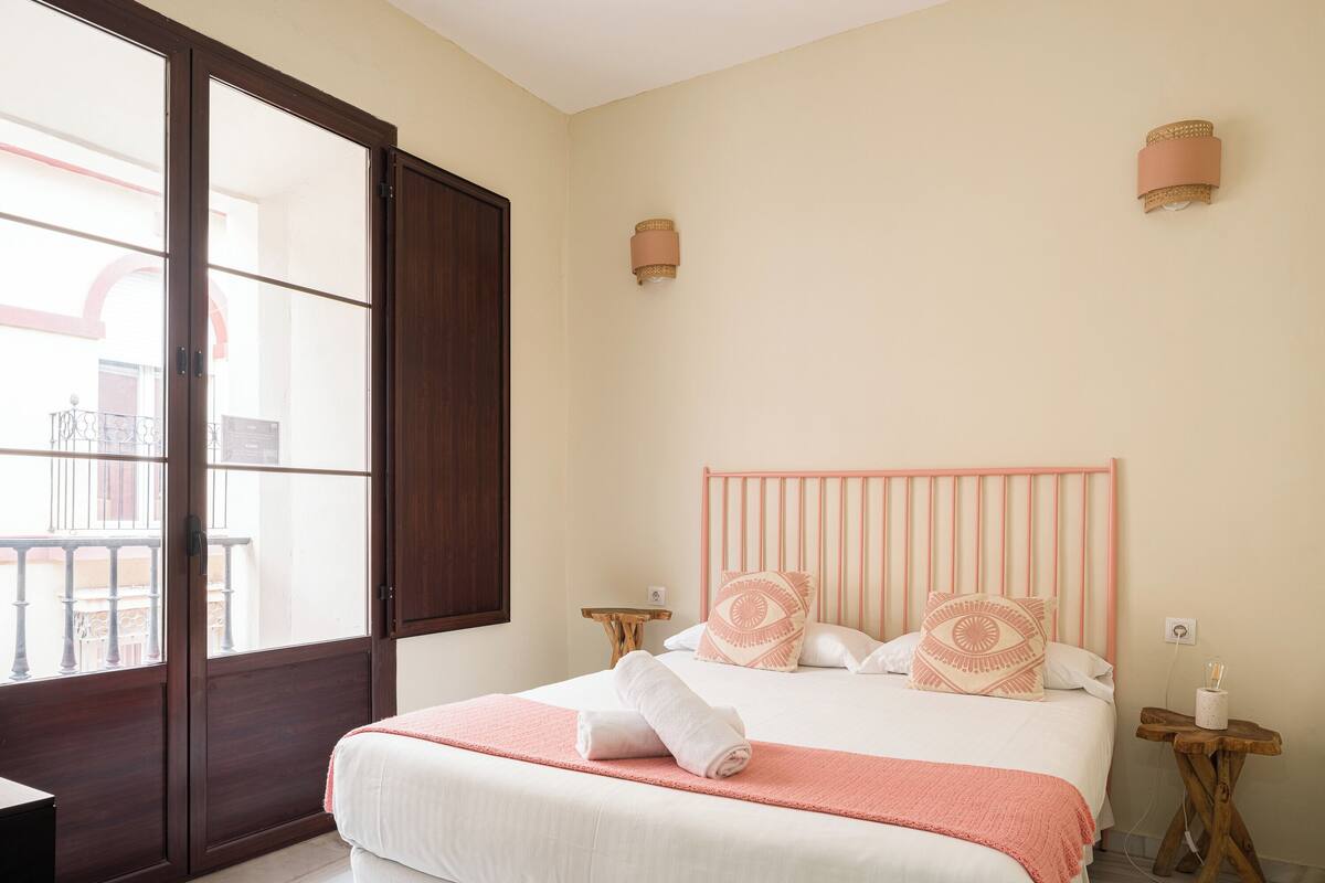 Un dormitorio con un ventanal grande, una cama con cabecera rosa y dos mesitas de noche.