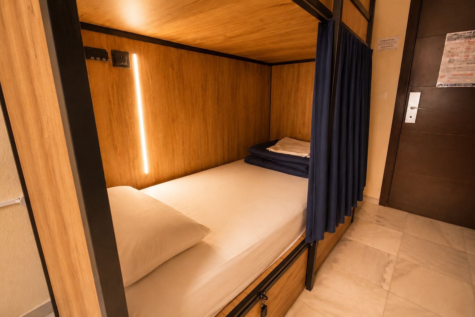 Habitación pequeña con cama individual, cabecero de madera, mesita de noche y cortina.