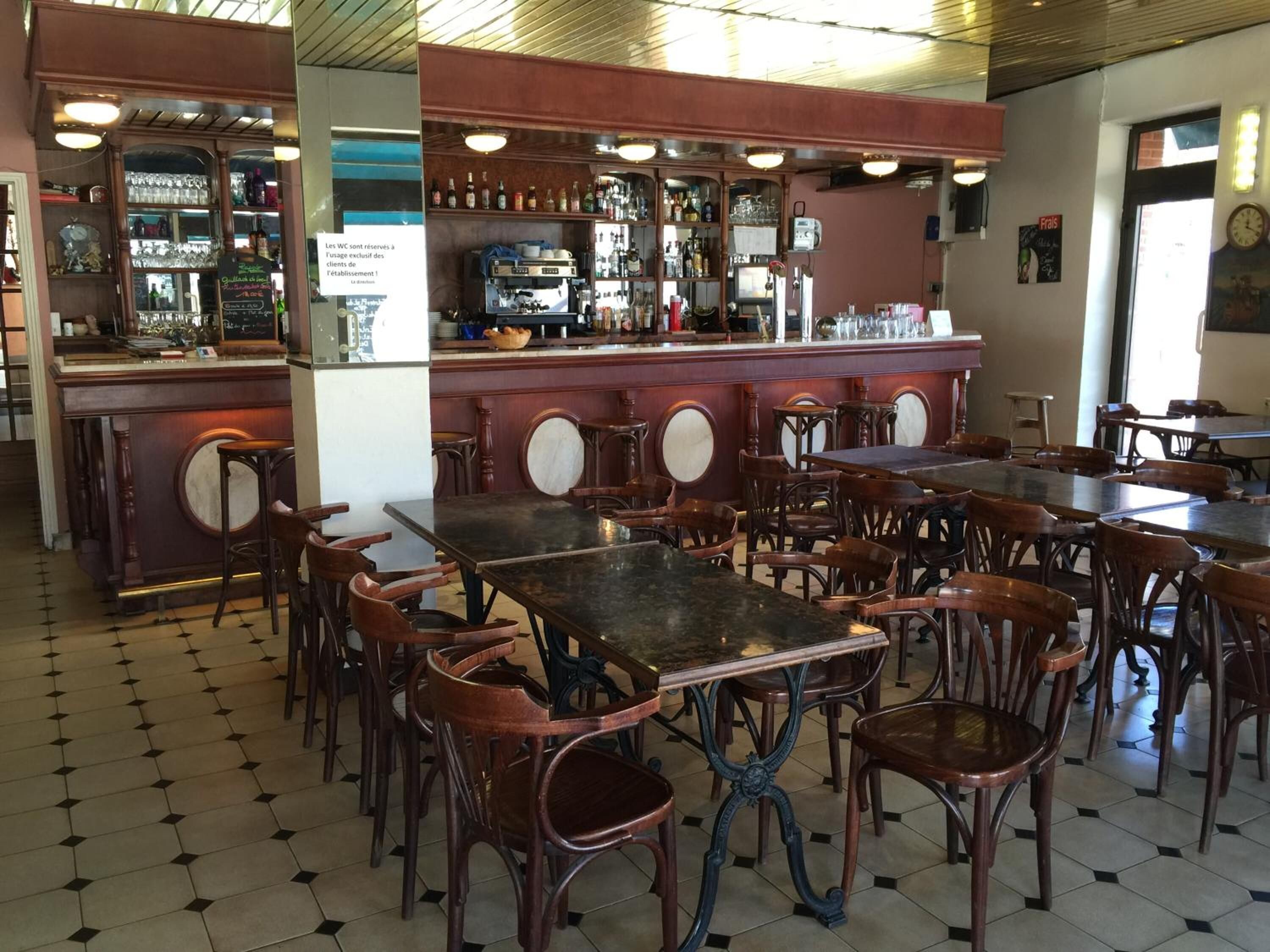 A restaurant interior with wooden tables and chairs, a bar area with shelves of bottles, and a tiled floor.