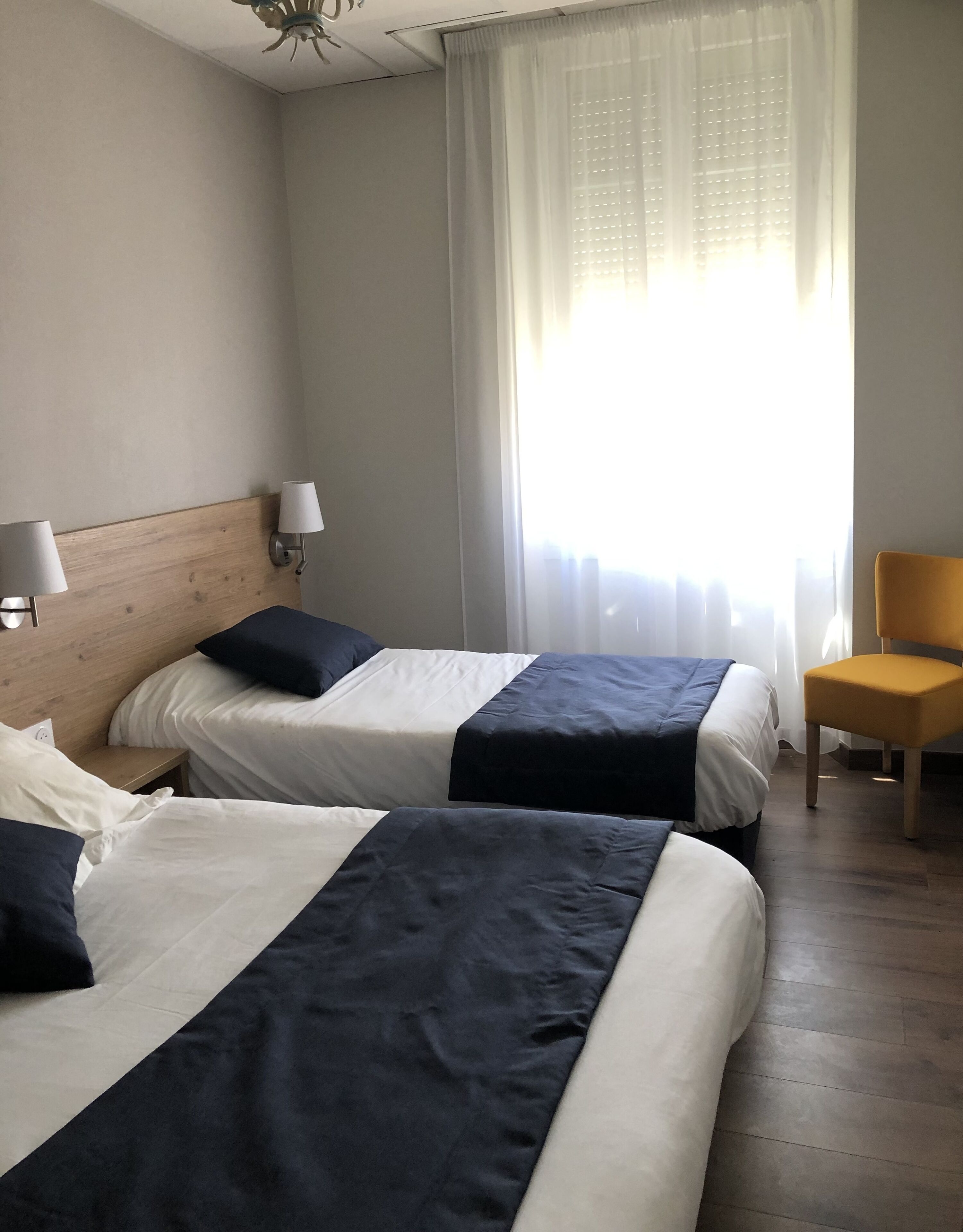 A hotel room with two beds, a chair, and a window with sheer curtains.